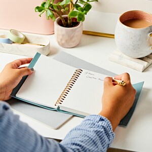 The A6 vegan leather notebook is laid open flat on a desk with a person writing on the inside. This notebook is open to reveal its cream blank pages.