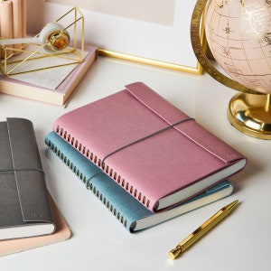 The a5 vegan leather notebook in dusky rose is stacked on top of a soft teal notebook. The notebooks are on a desk with a pink and gold globe of the world, a gold tape dispenser and a gold pen.