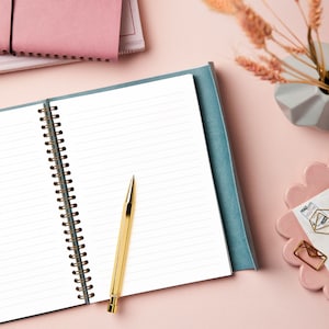 The A5 vegan leather notebook laid open on a soft peach background, with a soft teal cover. The inside of the notebook has lined paper and on top rests a gold pen