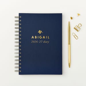 May include: A navy blue spiral-bound diary with gold lettering that reads "ABIGAIL 2026-27 diary." A gold pen, paper clips, and push pins are next to the diary on a white surface.