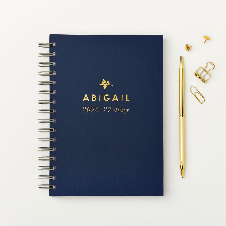 May include: A navy blue spiral-bound diary with gold lettering that reads "ABIGAIL 2026-27 diary." A gold pen, paper clips, and push pins are next to the diary on a white surface.