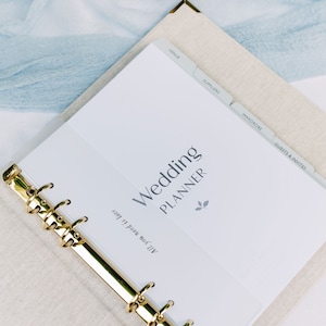 The wedding planner is laid open on a table to show the first page and a bookmark that features the phrase 'all you need is love'. The wedding planner is on a light blue linen covered table.