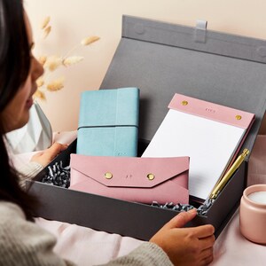 May include: A gray gift box with a blue leather journal, a pink leather notepad with the word "Lucy" on it, a pink leather envelope with the initials "A.P." on it, and a gold pen.