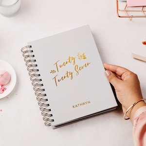 May include: A light gray spiral-bound notebook featuring gold foil text that reads Twenty Twenty Seven with decorative leaf accents and the name KATHRYN printed in gold below. The notebook has a metallic gold wire binding along the left side.