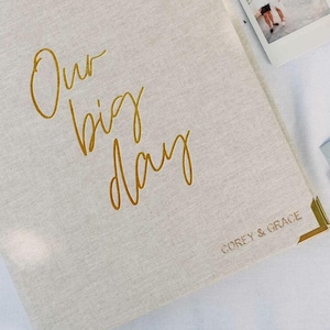 May include: A beige linen-covered photo album featuring the gold foil text Our big day in a cursive script on the front cover. The bottom right corner includes the gold foil text COREY & GRACE and a gold metal corner protector.