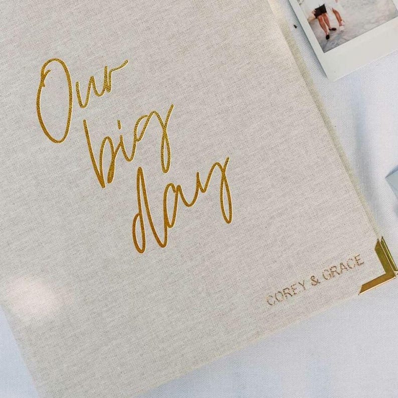 May include: A beige linen-covered photo album featuring the gold foil text Our big day in a cursive script on the front cover. The bottom right corner includes the gold foil text COREY & GRACE and a gold metal corner protector.