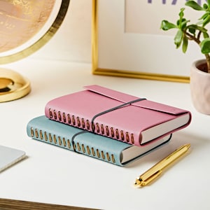 Two A6 vegan leather notebooks stacked on top of each other. The soft teal notebook is on the bottom and the dusky rose is on the top. The notebooks are on a white desk with a gold pen.