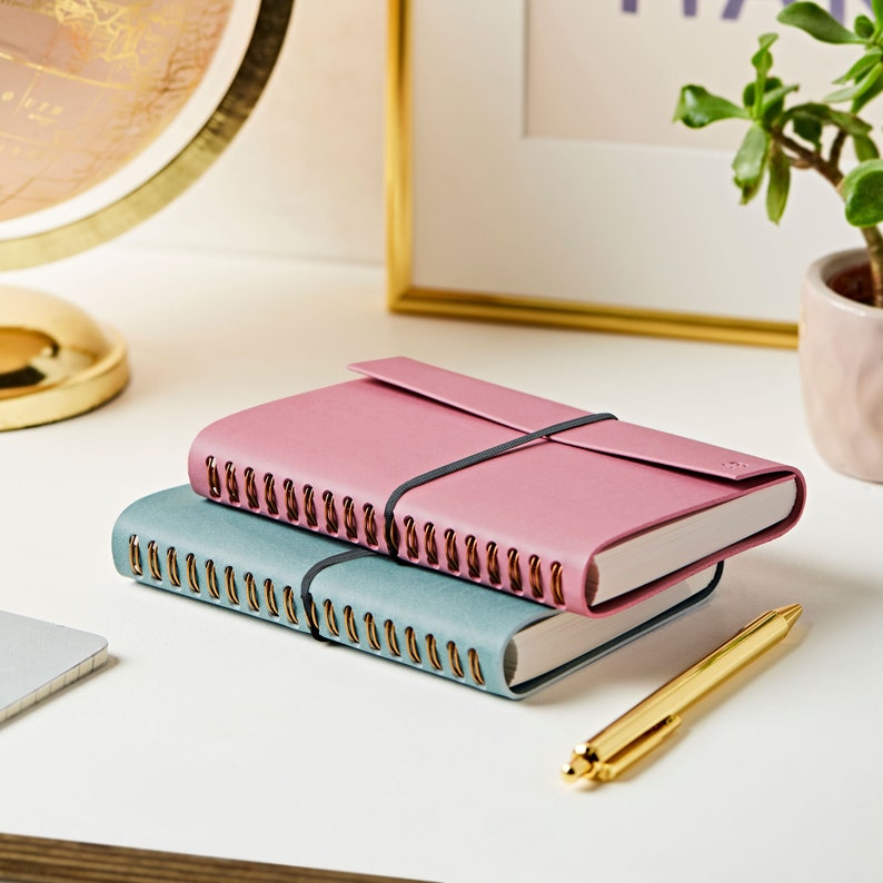 Two A6 vegan leather notebooks stacked on top of each other. The soft teal notebook is on the bottom and the dusky rose is on the top. The notebooks are on a white desk with a gold pen.