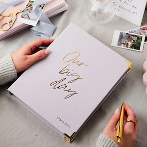 The lilac wedding planner features the phrase 'our big day' in a cursive font. The names SAM & KATY have been embossed in gold foil on the bottom right hand side of the planner. The lilac wedding planner is on a desk with polaroids.