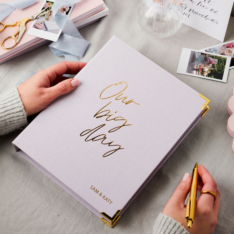 The lilac wedding planner features the phrase 'our big day' in a cursive font. The names SAM & KATY have been embossed in gold foil on the bottom right hand side of the planner. The lilac wedding planner is on a desk with polaroids.