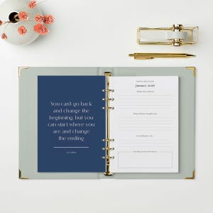 May include: Open journal with a blue page featuring a quote by C.S. Lewis and a white page with prompts for January 2026. Gold pen and tape dispenser are on the white surface. A vase with pink flowers is in the upper left corner.
