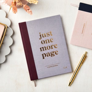 Könnte beinhalten: Ein graues und dunkelweinrotes Hardcover-Buchjournal mit goldener Folienprägung auf dem Einband, das "just one more page" und "A BOOK JOURNAL" sowie die Initialen "CLB" unten rechts zeigt, begleitet von einem goldenen Metallstift.