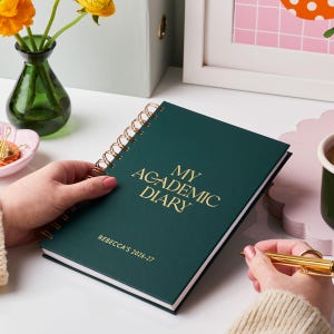 May include: A dark green academic diary with a gold spiral binding and gold lettering that reads "MY ACADEMIC DIARY". The diary also has the name "REBECCA'S 2026-27" printed on the cover. A gold pen is held near the open diary.