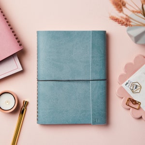 The personalised A5 vegan leather notebook in soft teal, personalised in the bottom right hand corner with the initials SB. The notebook is next to a gold pen and a candle and some paperclips.