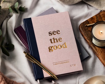 May include: A pink gratitude journal with a navy blue spine and gold lettering that reads "see the good". The journal is open and a gold pen is lying on top of it.
