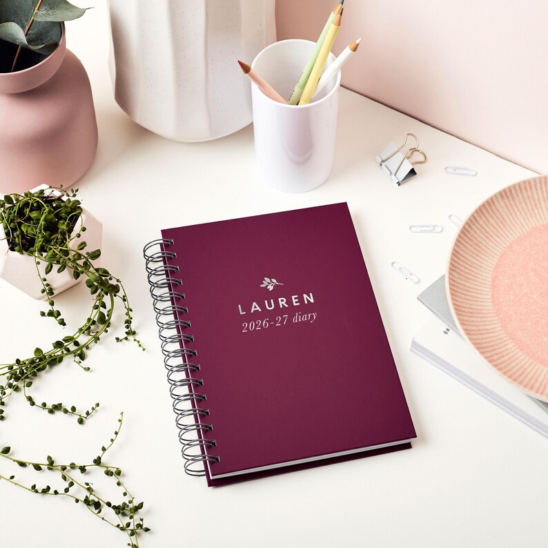 May include: A burgundy spiral-bound diary with the name "LAUREN" and the dates "2026-27 diary" printed in white. The diary is on a white surface, with pencils, a small plant, and a decorative plate nearby.
