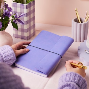 The a5 vegan leather notebook in lilac being held by a woman, personalised with the initials CDR in the bottom right hand corner. The lilac notebook is on a desk with a lilac pen pot and a lilac flower pot.