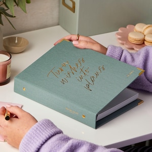 May include: A teal-colored planner with gold lettering that reads "Turn wishes into plans." The planner has gold corner protectors and a matching spine. The planner is open, revealing blank pages. A person is holding the planner.