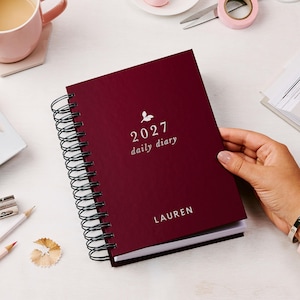 May include: A dark burgundy spiral-bound daily diary with a black wire coil. The cover features a small white leaf icon above the text 2027 daily diary and the name LAUREN printed in white at the bottom center of the front cover.