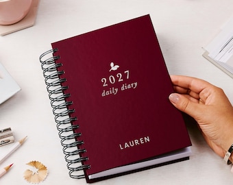 May include: A dark burgundy spiral-bound daily diary with a black wire coil. The cover features a small white leaf icon above the text 2027 daily diary and the name LAUREN printed in white at the bottom center of the front cover.