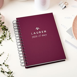 May include: A burgundy spiral-bound diary with the name "LAUREN" and the dates "2026-27 diary" printed in silver. The diary is on a white surface with other office supplies and greenery in the background.