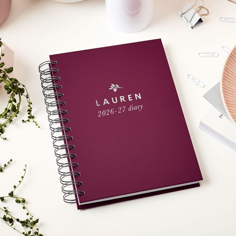 May include: A burgundy spiral-bound diary with the name "LAUREN" and the dates "2026-27 diary" printed in silver. The diary is on a white surface with other office supplies and greenery in the background.