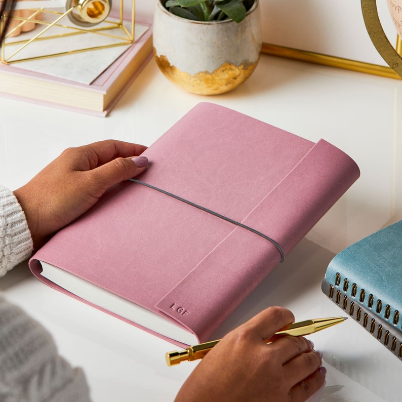 A person holding the personalised A5 vegan leather notebook in dusky pink. The notebook features a dark grey elastic cord to secure the notebook. Personalised in the bottom right hand corner with the initials LGF.