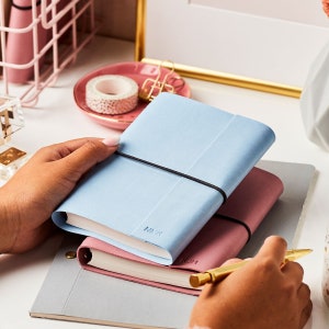 The A6 vegan leather notebook in powder blue, stacked on top of a pink notebook with a grey band around the middle. The notebook is personalised with the initials NMK. The blue notebook is being held by a pair of hands at a desk.