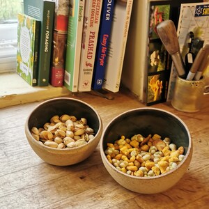以下が含まれることがあります： 内側が濃い緑色の、小さくて丸いベージュ色の陶器のボウルが2つ。1つのボウルにはピスタチオが、もう1つにはナッツとスナックのミックスが入っています。ボウルは木製の表面に置かれ、背景には料理本とペン立てがあります。