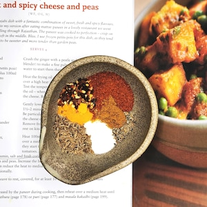 May include: A speckled, oval-shaped bowl containing a variety of spices, including cumin seeds, turmeric, and chilli powder. The bowl is part of a recipe for a dish with cheese and peas. The recipe is on a page with text.