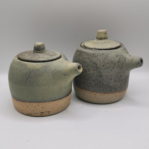 May include: Two ceramic teapots with lids. The teapots are round with a spout and a handle on top. The teapots are speckled gray and green with a tan base. The teapots are handmade and have a rustic look.