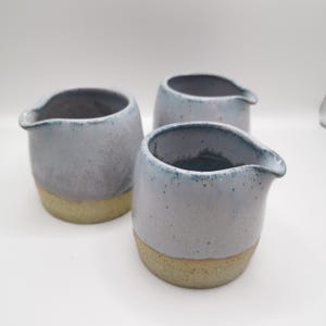 Puede incluir: Tres jarras de cerámica hechas a mano. Las jarras son de color azul claro con una textura moteada y una base beige. Cada jarra tiene un pico para verter. Las jarras están dispuestas sobre una superficie blanca.
