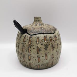 以下が含まれることがあります： 蓋と小さな黒いスプーンが付いたセラミック製の砂糖入れ。ボウルは、縦のオレンジ色のダッシュと濃い茶色の縁が付いた、斑点のあるオリーブグリーン色です。スプーンはボウルの側面にあります。