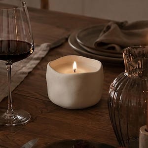 May include: A white ceramic candle with a wavy, organic rim and a lit wick, placed on a wooden table alongside a wine glass filled with red wine, a bowl of red grapes, two pomegranates, a glass vase with a brown taper candle, and dinnerware.