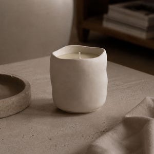 Puede incluir: Una vela de color crema en un recipiente de cerámica texturizado y redondeado. La vela está sobre una superficie de tonos neutros, con un plato pequeño y un paño doblado cerca. La vela tiene una sola mecha.