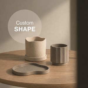 May include: A set of three concrete items including a beige planter with a wavy rim, a smaller grey fluted planter, and a grey irregular cloud-shaped tray. The text Custom SHAPE is visible in a circle above the beige planter on the left side.