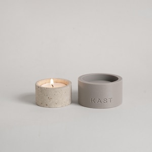 Peut inclure: Deux petits bougeoirs cylindriques. L'un est allumé avec une petite flamme, l'autre est vide. Les bougeoirs sont gris et en béton. Le mot "KAST" est imprimé sur le côté du bougeoir vide.