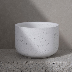 May include: A white speckled planter with a rounded base and a wide opening. The planter is made of a concrete-like material and sits on a gray surface. The speckled design features small black dots throughout the white surface.