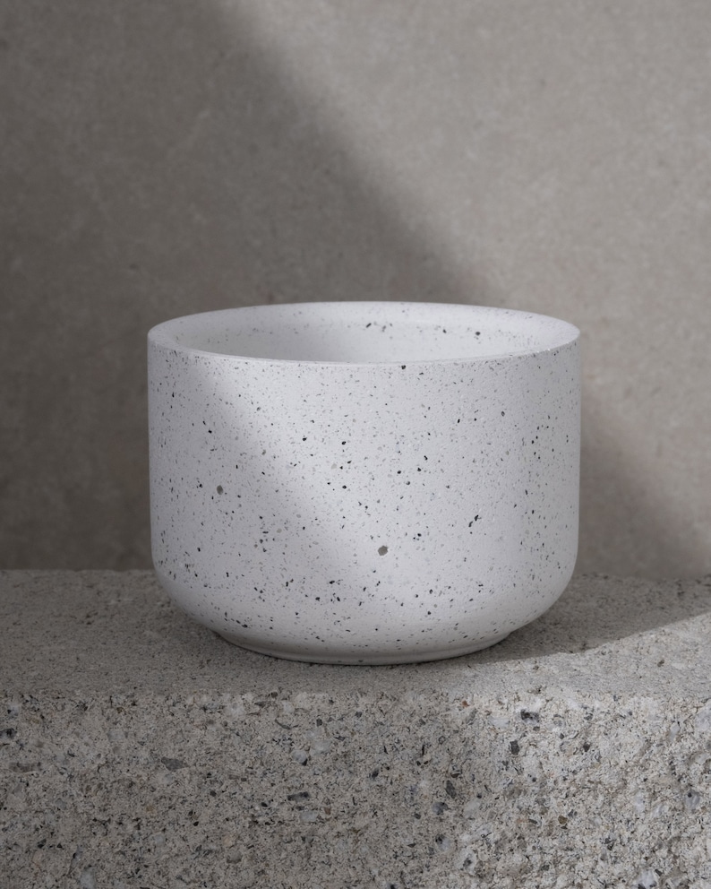 May include: A white speckled planter with a rounded base and a wide opening. The planter is made of a concrete-like material and sits on a gray surface. The speckled design features small black dots throughout the white surface.