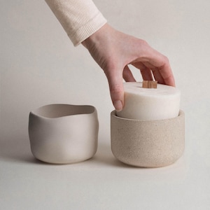 Puede incluir: Una mano coloca una vela blanca con una mecha de madera en un soporte de cerámica beige moteado. A la izquierda hay otro recipiente de cerámica beige claro, liso y vacío. El fondo es de un blanco roto neutro.