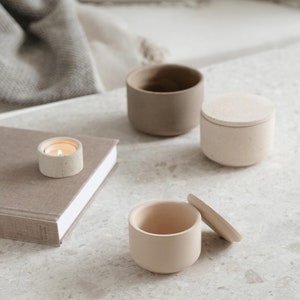 May include: A collection of neutral-toned ceramic containers and a small candle. The containers include a small open cup, a lidded jar, and a larger open cup. The candle is lit, casting a warm glow. The items are arranged on a speckled surface.