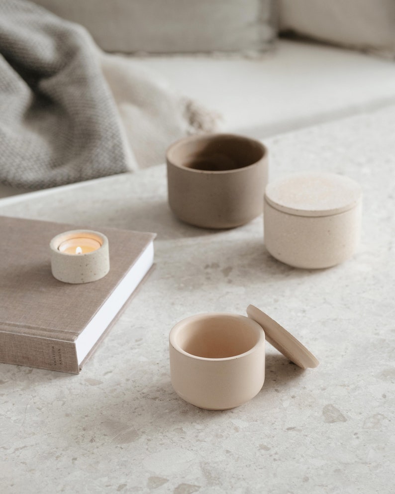 May include: A collection of neutral-toned ceramic containers and a small candle. The containers include a small open cup, a lidded jar, and a larger open cup. The candle is lit, casting a warm glow. The items are arranged on a speckled surface.