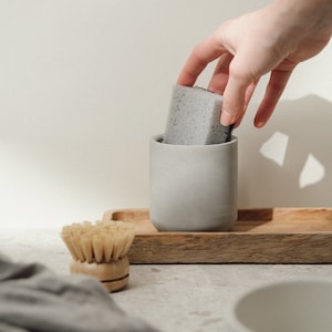 May include: A grey ceramic dish soap dispenser with a grey sponge inside. The dispenser is sitting on a wooden tray with a wooden brush next to it.