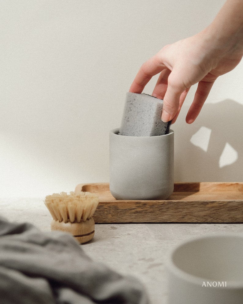 May include: A grey ceramic dish soap dispenser with a grey sponge inside. The dispenser is sitting on a wooden tray with a wooden brush next to it.