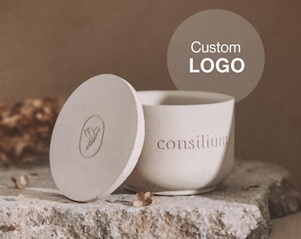 May include: A light beige cylindrical container with a matching circular lid. The side of the container features the embossed text 'consilium', while the lid displays a small circular emblem with a leaf-like design. The surface has a matte texture.
