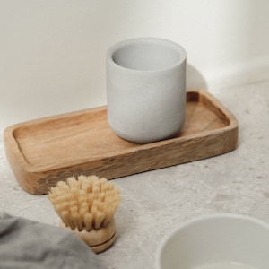 May include: A grey ceramic cup sits on a wooden tray with a natural wood finish. A small wooden brush with natural bristles is in the foreground. The background is a light grey textured surface.