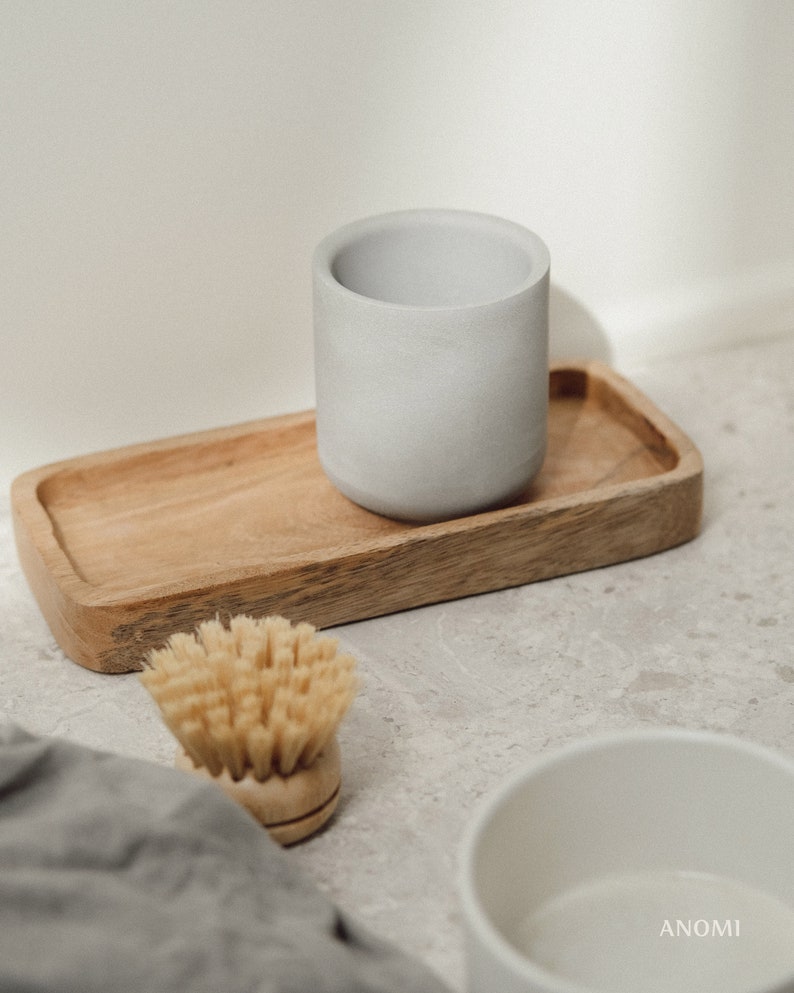 May include: A grey ceramic cup sits on a wooden tray with a natural wood finish. A small wooden brush with natural bristles is in the foreground. The background is a light grey textured surface.
