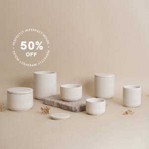 Może przedstawiać: Zestaw sześciu białych ceramicznych słoików na świece z pokrywkami, z okrągłą naklejką z napisem "Perfectly Imperfect Molds 50% Off".