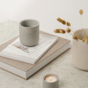 May include: A grey concrete candle holder with a lit tea light on a white countertop. A stack of books with a grey concrete cup on top and a vase with dried flowers are in the background.