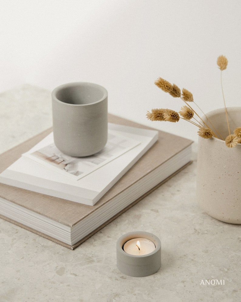 May include: A grey concrete candle holder with a lit tea light on a white countertop. A stack of books with a grey concrete cup on top and a vase with dried flowers are in the background.